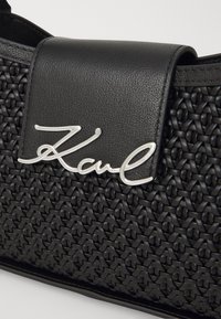 Borsa a tracolla in tessuto nero con un cappuccio in pelle liscia, caratterizzata da un accento in logo argentato. La texture è intricatata con un design strutturato.
