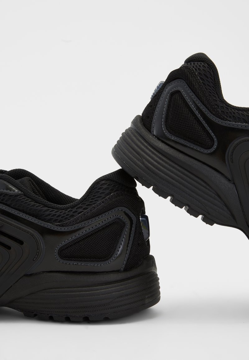 Paire de baskets de sport noires avec mesh texturé et semelles en caoutchouc sur fond gris clair, vues de dos et de profil.