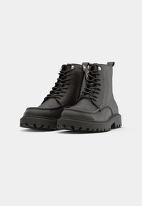 Svarta läderkampskängor med snörning, sidodragkedjor, kraftiga gummisålar och texturerade detaljer på ovandelen och sulan.