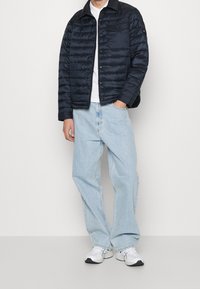 Mężczyzna w czarnej pikowanej kurtce, białej koszulce, jasnoniebieskich luźnych dżinsach i białych sneakersach, stojący z jedną ręką w kieszeni.