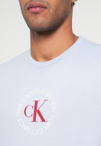 Jasnoniebieska bawełniana koszulka z okrągłym nadrukiem zawierającym czerwone logo "cK" i biały tekst. Gładka faktura i klasyczny dekolt w stylu crew neck.