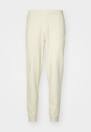 Un pantalon de survêtement couleur crème en tissu doux avec une taille élastique et des poignets élastiques. Il présente un petit écusson logo sur la hanche gauche.