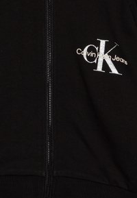 Črna kapuca z zadrgo, ki ima srebrno vezeno logo "Calvin Klein Jeans" in rebraste manšete na robu.