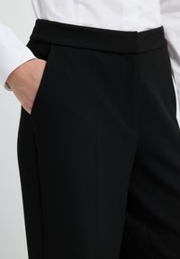 Persoon in zwarte op maat gemaakte trousers met hand in de linkerzak en een witte blouse met lange mouwen gestoken.