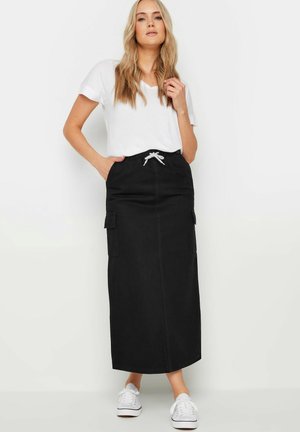 Falda maxi de mezclilla negra con bolsillos laterales, cintura con cordón y costuras blancas. Combinada con una camiseta blanca sencilla y zapatillas blancas.