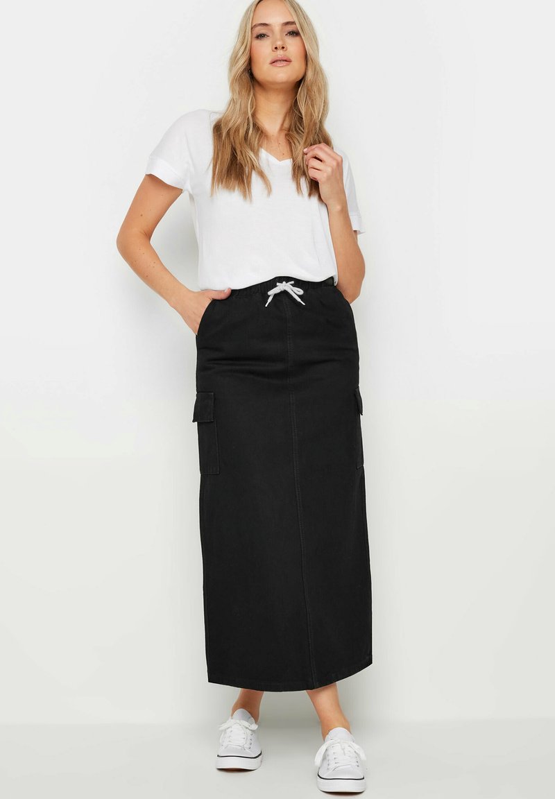 Maxi gonna in denim nero con tasche laterali, vita regolabile con cordoncino e cuciture bianche. Abbinata a una maglietta bianca semplice e sneakers bianche.
