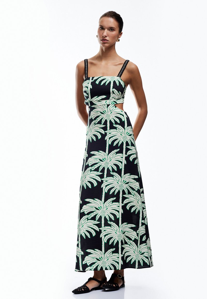 Schwarzes Maxikleid mit grünem Palmbaumdruck, Ausschnittdesign an der Taille und schmalen Trägern, die mit dekorativem Zubehör geschmückt sind.