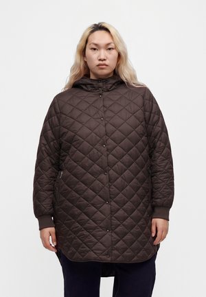 Personne aux longs cheveux blonds portant une longue veste matelassée marron avec des boutons-pression et des poches latérales zippées sur fond blanc.