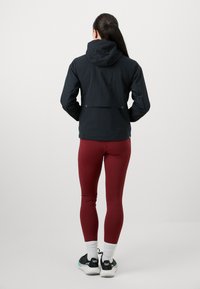 Črna kapucar z gladko teksturo, ki ima zadnji ventil in minimalističen dizajn. Obuta v maroon legice in črne teniske s teal akcenti.