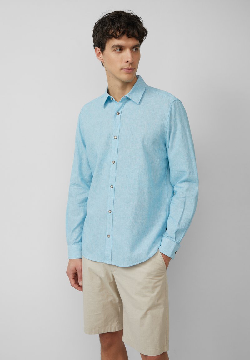 Hellblaues gewebtes Langarmhemd mit Button-Down-Kragen, subtiler Textur und Knopfdetails, kombiniert mit beigen Shorts.