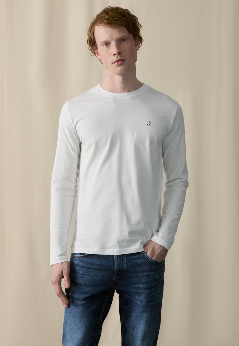 Hvid langærmet t-shirt lavet af bomuld, med rund hals og et lille broderet logo på venstre bryst, båret med blå jeans.
