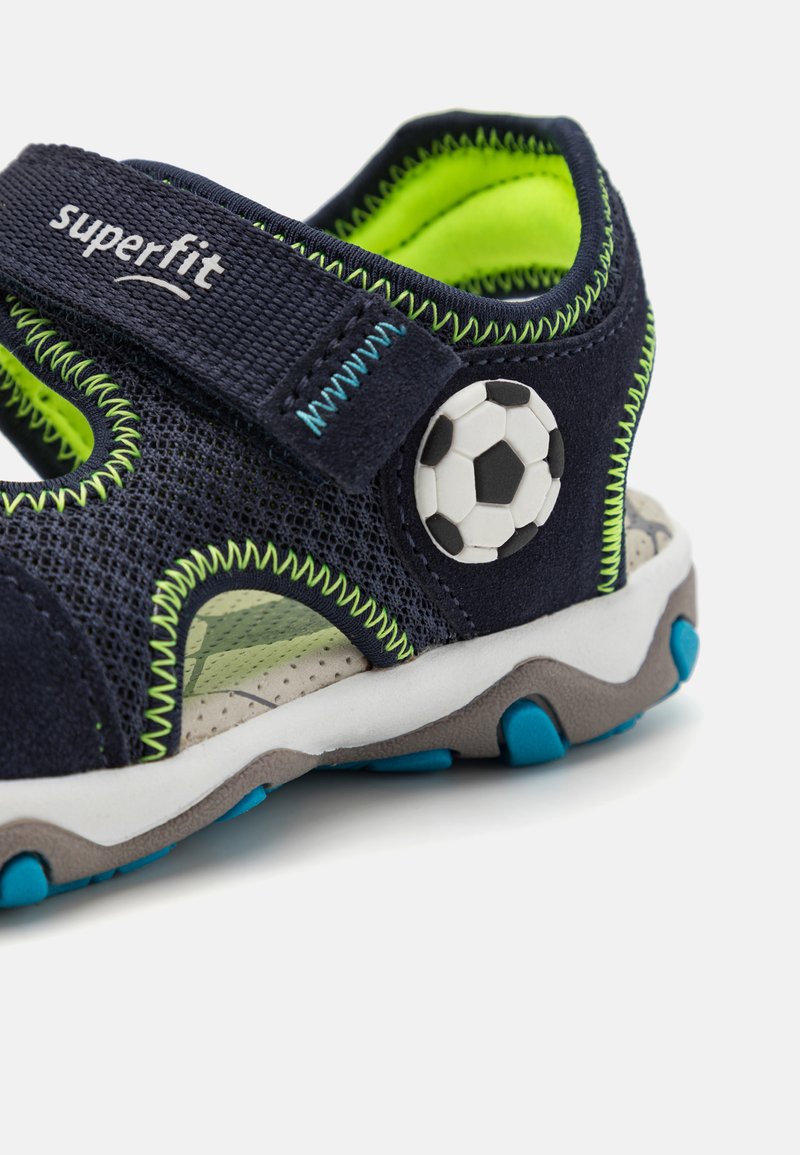Navyblaue Sandale mit Obermaterial aus Mesh und grünen Akzenten, mit einem Fußball-Detail, verstellbarem Riemen und grauer Gummisohle mit blauen Profilen.