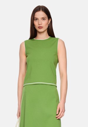 Frau mit langen braunen Haaren trägt ein ärmelloses grünes Oberteil und einen passenden grünen Rock mit weißem Besatz vor hellem Hintergrund.