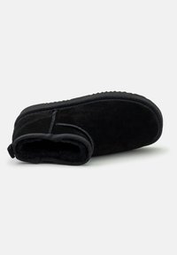 Zapatilla de ante negro con un suave forro de pelaje, un perfil bajo y una suela de goma texturizada. Presenta costuras a lo largo de las costuras y una pestaña en el talón.