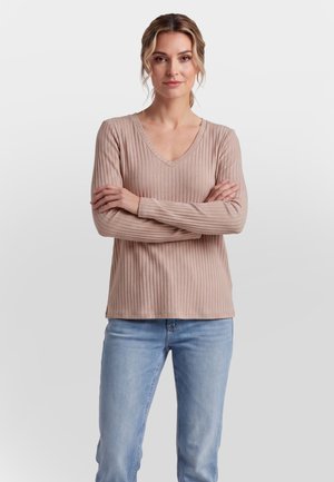 Ung kvinde med blondt hår iført en beige ribstrikket V-hals sweater og lyseblå jeans, står med armene over kors mod en ensfarvet baggrund.