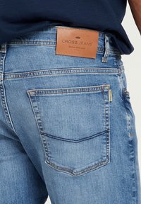 Lichtblauwe denim jeans met een leren taillebandlabel met "CROSS JEANS" en twee achterzakken met subtiele stikdetails.