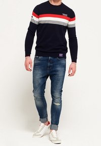 Námořnický pletený svetr s červenými, šedými a bílými pruhy, kombinovaný s opotřebovanými modrými džínami a bílými teniskami s červenými detaily.