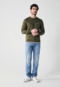 Olijfgroene sweatshirt met geribbelde manchetten en zoom, gecombineerd met lichtblauwe jeans en witte sneakers. Model staat met gekruiste armen, neutrale achtergrond.