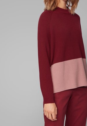Femme portant un pull en tricot à manches longues rouge foncé avec une large bande rose clair, assorti à un pantalon rouge foncé sur fond gris.