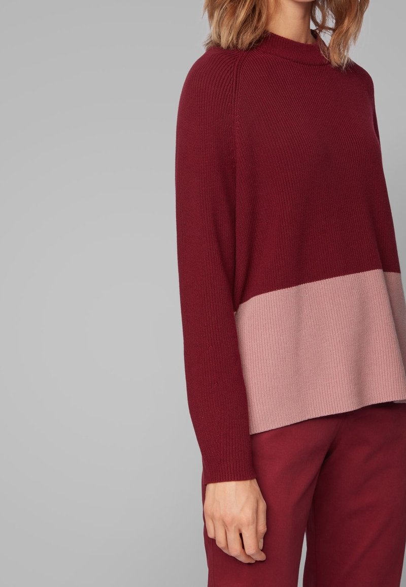 Femme portant un pull en tricot à manches longues rouge foncé avec une large bande rose clair, assorti à un pantalon rouge foncé sur fond gris.