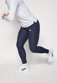 Mørkeblå treningslegger med en glatt tekstur, med en hvit Adidas-logo på venstre lår, sammen med hvite treningssko.