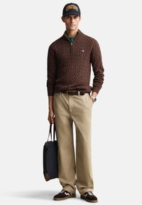 Bruine kabelgebreide trui, groene overhemd met kraag, kaki broek, bruine schoenen, navy tas en navy pet met gele tekst. Alle items in een casual pasvorm.