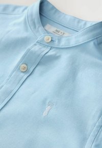 Hellblaues Poloshirt aus weichem Stoff mit kurzem Kragen und zwei weißen Knöpfen, verziert mit einer kleinen weißen Hummerstickerei auf der Brust.