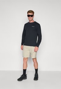 Chemise à manches longues noire, shorts kaki, baskets noires et lunettes de soleil. Le tissu est en coton ; les couleurs sont unies. Logo visible sur la zone de la poitrine.