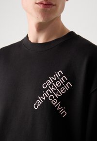 Sweatshirt negru din bumbac, cu un aranjament diagonal al textului "calvin" în roz deschis pe partea stângă. Guler rotund și textură netedă.