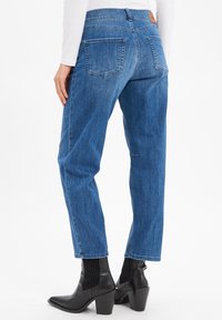 Femme portant un jean droit bleu et des bottes à talons noires, vue de dos contre un fond blanc.