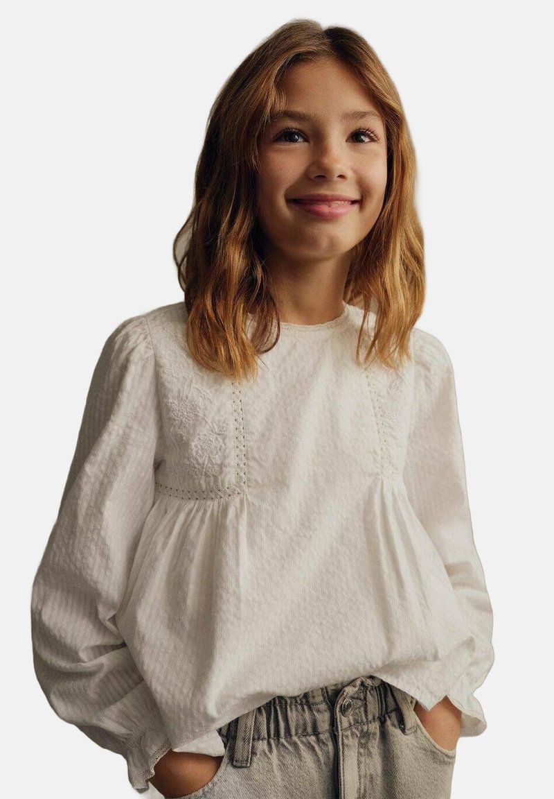 Blusa bianca a maniche lunghe in tessuto testurizzato, con dettagli in pizzo e vita arricciata, abbinata a pantaloni grigi chiari a vita alta.