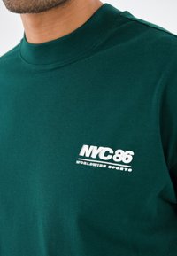 Grünes Baumwoll-T-Shirt mit Rundhalsausschnitt, geripptem Kragen und bedrucktem "NYC-86"-Logo sowie dem Text "WORLDWIDE SPORTS" in Weiß auf der Vorderseite.