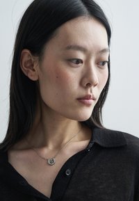 Ασημένιο κολιέ με μενταγιόν σε σχήμα πετάλου, φορεμένο πάνω σε μαύρο τοπ με γιακά και υφή. Απλός σχεδιασμός, λείο φινίρισμα και ευαίσθητη αλυσίδα.