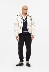 Witte pufferjack met capuchon en voorrits, gecombineerd met een marineblauwe gebreide trui, zwarte sweatpants en zwarte instappers met hardware accenten.