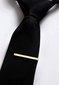 Pince à cravate en or sur une cravate en satin noir. La cravate est lisse avec une légère brillance, tandis que la pince est une barre rectangulaire aux lignes épurées.