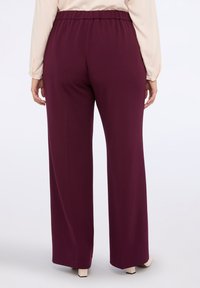 Woman wearing high-waist, wide-leg burgundy pants and a long-sleeve beige blouse, shown from the back against a white background.