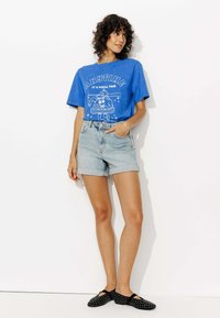 Femme aux cheveux bouclés se tient contre un mur blanc portant un T-shirt graphique bleu "C'est toujours l'heure de la pizza", un short en denim clair et des chaussures plates noires à clous.