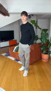 Fiatal férfi fekete pulóverben, bézs katonazsebes nadrágban és fehér sportcipőben áll egy nappaliban, ahol barna kanapé, nagy cserepes növény és televízió található.