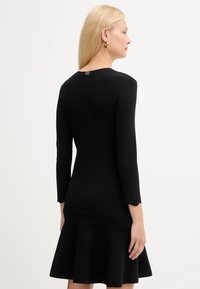 Donna con lunghi capelli biondi che indossa un vestito nero aderente con bordi ondulati, orecchini a cerchio dorati, rivolta di lato su uno sfondo neutro.