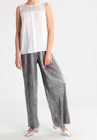 Top bianco senza maniche con dettagli in pizzo e una patta, abbinato a pantaloni larghi plissettati argento e scarpe metallizzate coordinate.