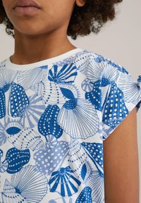 La camisa de manga corta presenta un fondo blanco con patrones de conchas azules y lunares, hecha de suave tela de algodón. Los acentos incluyen detalles fruncidos en los hombros.