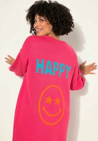 Un pull surdimensionné rose présente le mot "HAPPY" en bleu canard et un visage souriant avec des yeux en forme d'étoile orange au dos. Texture douce et design décontracté.