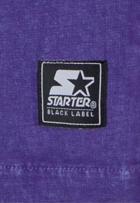 Tissu violet avec une finition texturée présentant une pi�èce rectangulaire noire sur laquelle est inscrit "STARTER® BLACK LABEL" en lettres blanches.