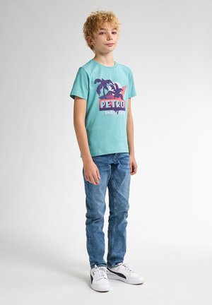 Turkise t-shirt med palme grafik og teksten "PETRO INDUSTRIES", sammen med blå jeans og hvide sneakers, stående foran en lys baggrund.