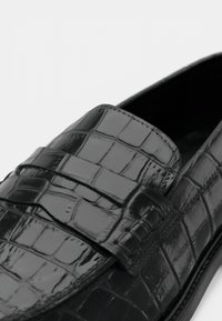 Čierne kožené mokasíny s krokodíliu textúrou, s širokým remienkom cez hornú časť a jemným prešívaním pozdĺž okrajov.