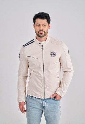 Beige jas met een hoge kraag, ritssluiting, schouderstrepen en een geborduurd embleem. Voorzien van zijritssluitingen en een gladde textuur.