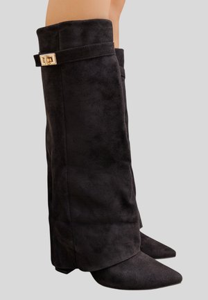 Bottes hautes en daim noir, avec bouts pointus, talons compensés et sangles à boucle dorée près du haut, portées sur des jambes nues.