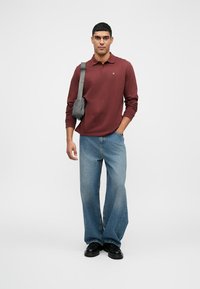 Polo shirt bordó cu mânecă lungă, blugi largi bleu, pantofi negri și o geantă gri tip crossbody. Modelul stă în fața unui fundal alb simplu.