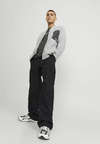 Giacca bomber grigia, pantaloni cargo neri con tasche multiple, t-shirt grigia e sneakers bianche con accenti neri. Sfondo semplice.