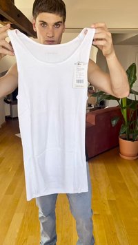 White ribbed tank top with wide neckline and tag attached, displayed by a person. Background includes indoor plants and a sofa.